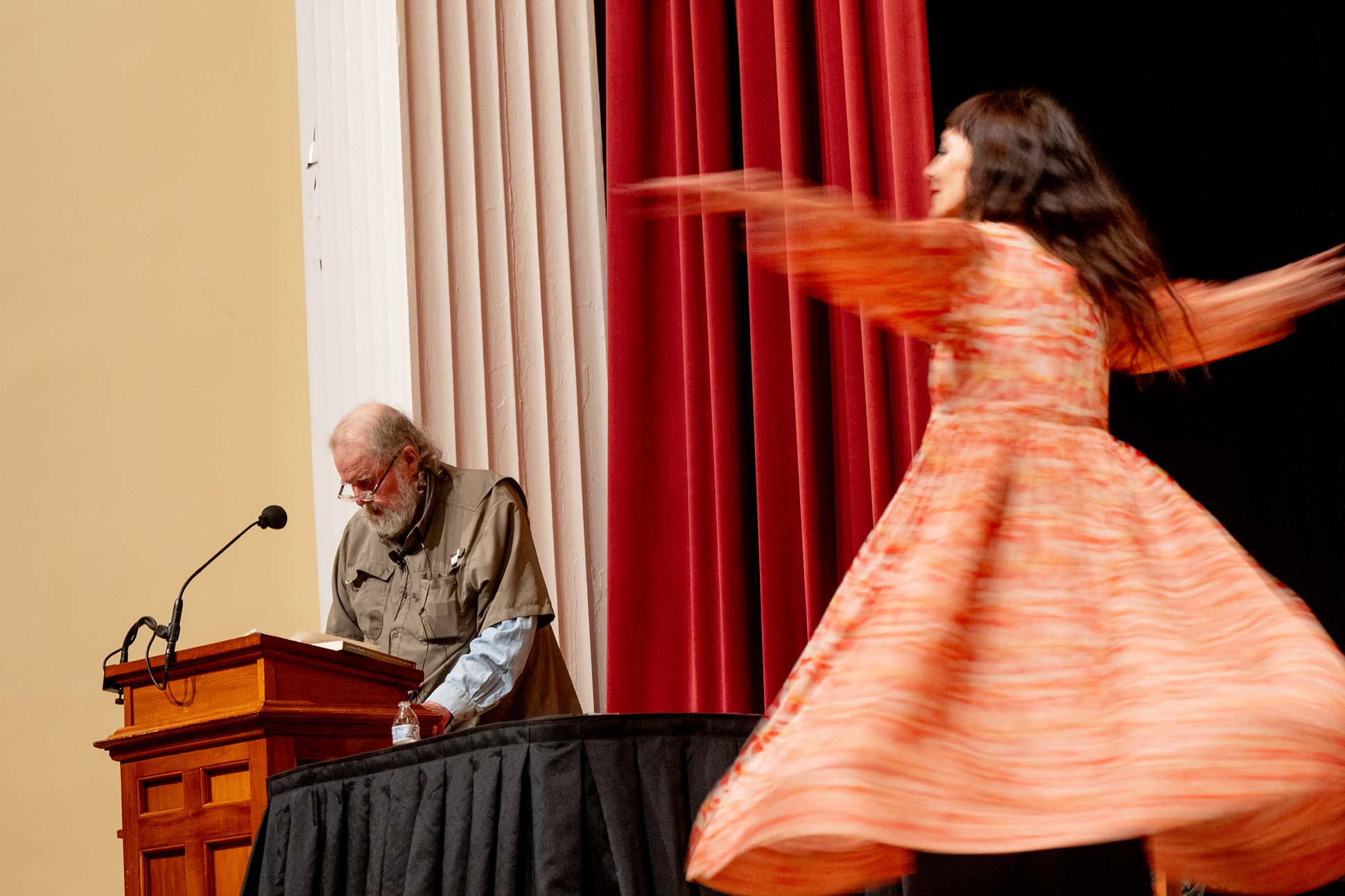 A man stands at a podium reading during the Willson Center event, while a woman in a patterned dress twirls nearby on stage with red curtains in the background, highlighting moments featured in the 2025 Annual Report.