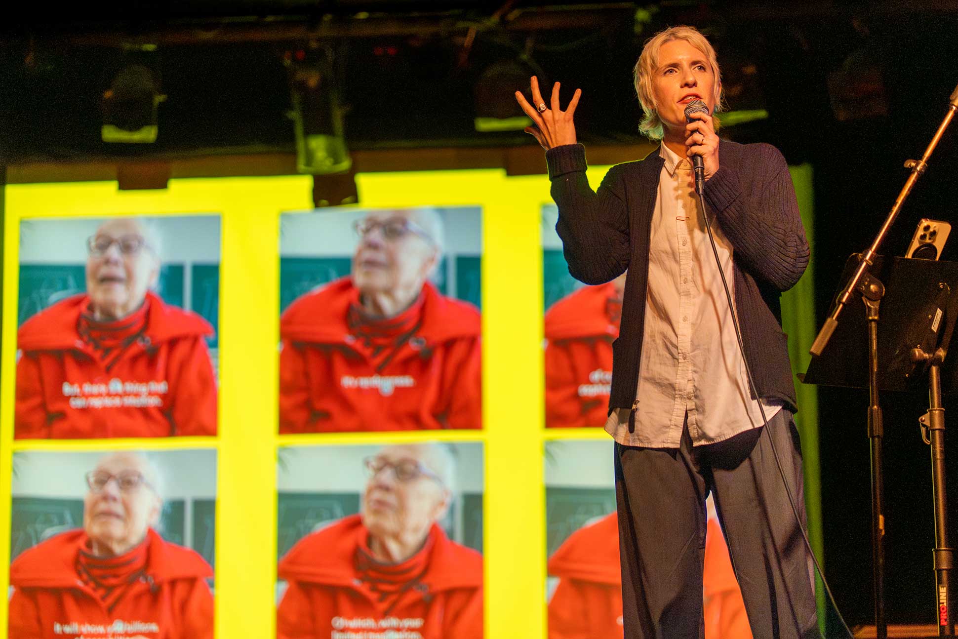 A person speaks into a microphone on stage, gesturing with one hand, with a large screen behind them displaying repeated images of an elderly person in a red jacket.