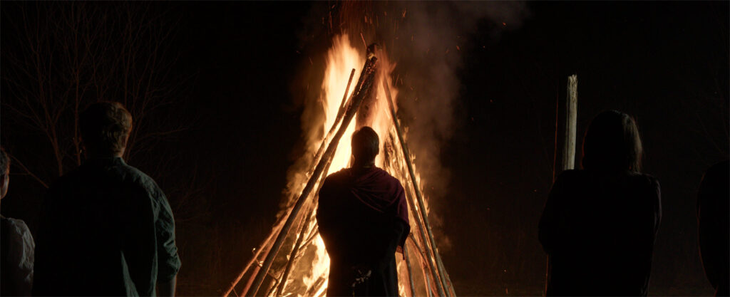 Four people stand outdoors at night, facing a large bonfire made of upright wooden logs, flames and smoke rising into the dark sky as they celebrate their Graduate Research Awards.