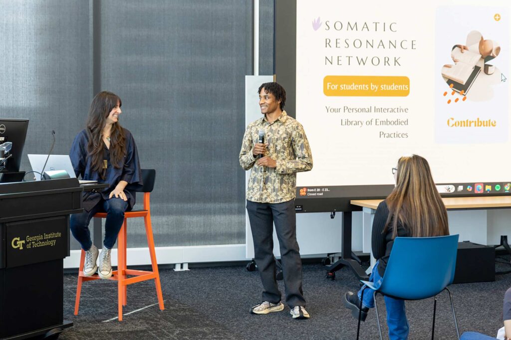 A man speaks into a microphone while standing near a seated woman; a large screen behind them displays the Somatic Resonance Network website at Georgia Institute of Technology.