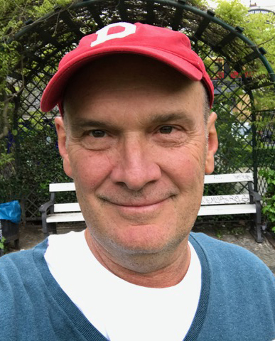 A man wearing a red cap and blue sweater is smiling outdoors in front of a white bench and garden trellis, capturing the welcoming spirit of the 2025 Willson Center Annual Report.